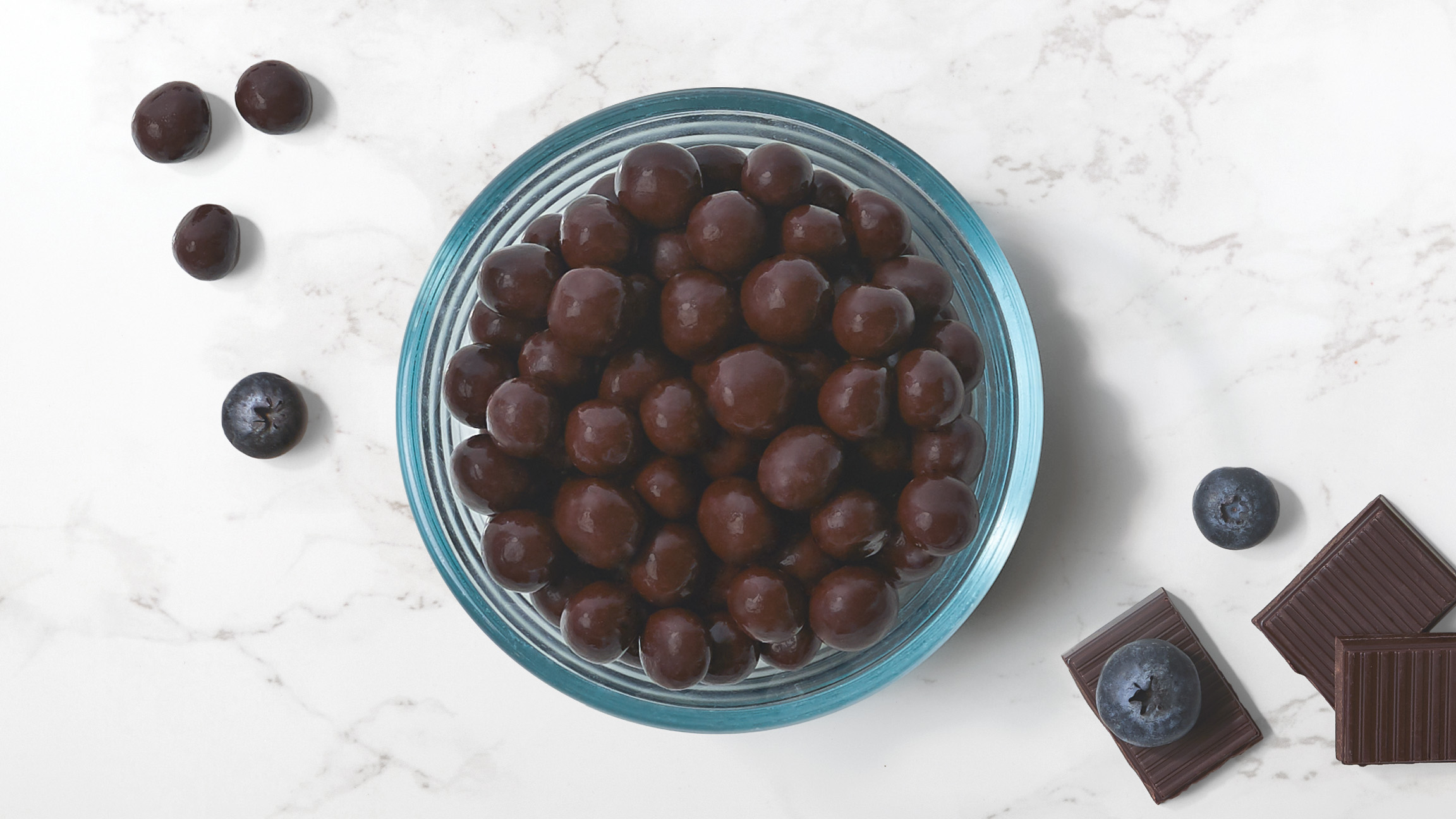 Dark chocolate blueberries in a glass bowl with scattered fresh blueberries and pieces of chocolate