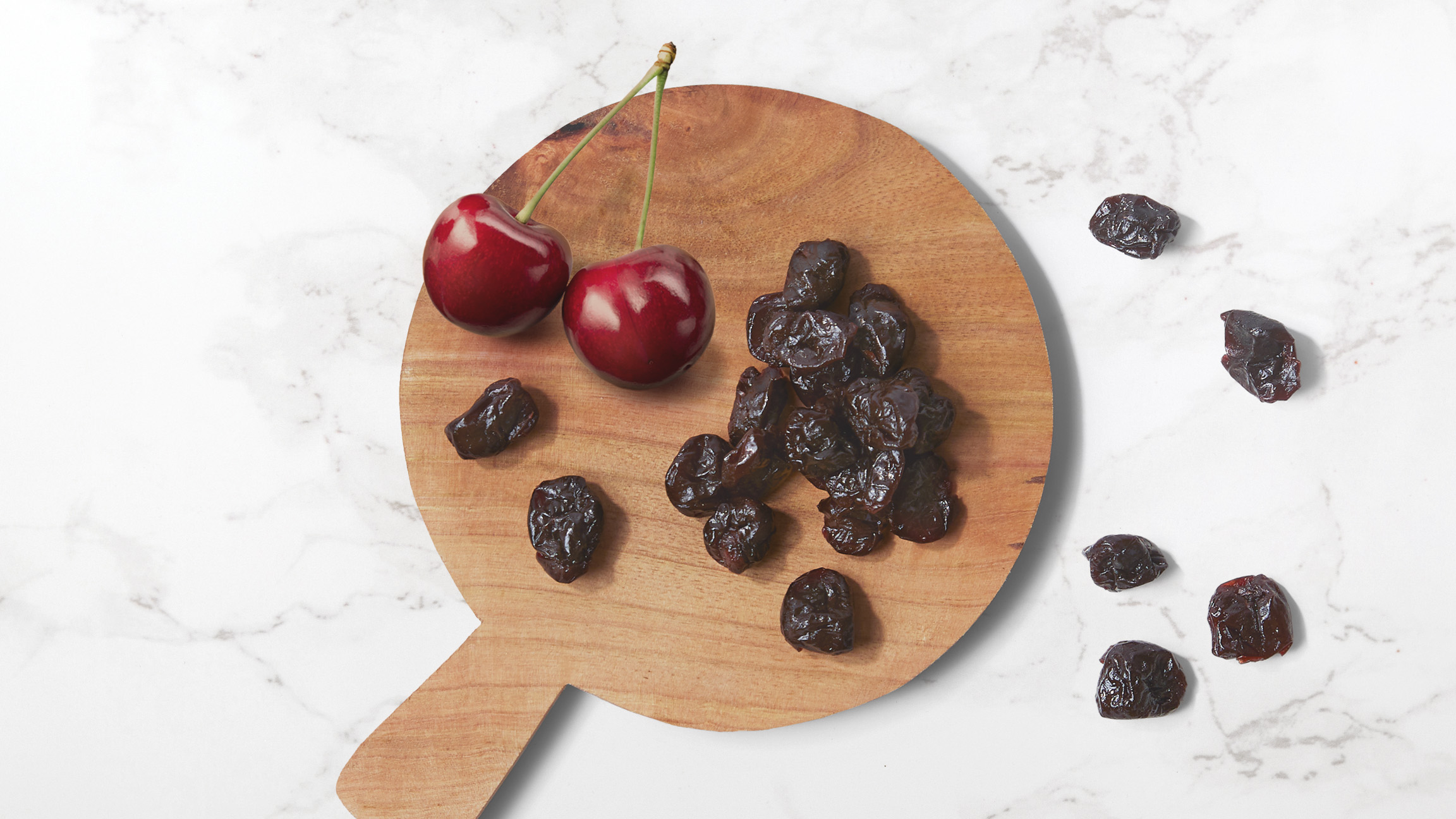 Dried cherries on a circular wood serving platter with dried and fresh cherries scattered on a marble counter