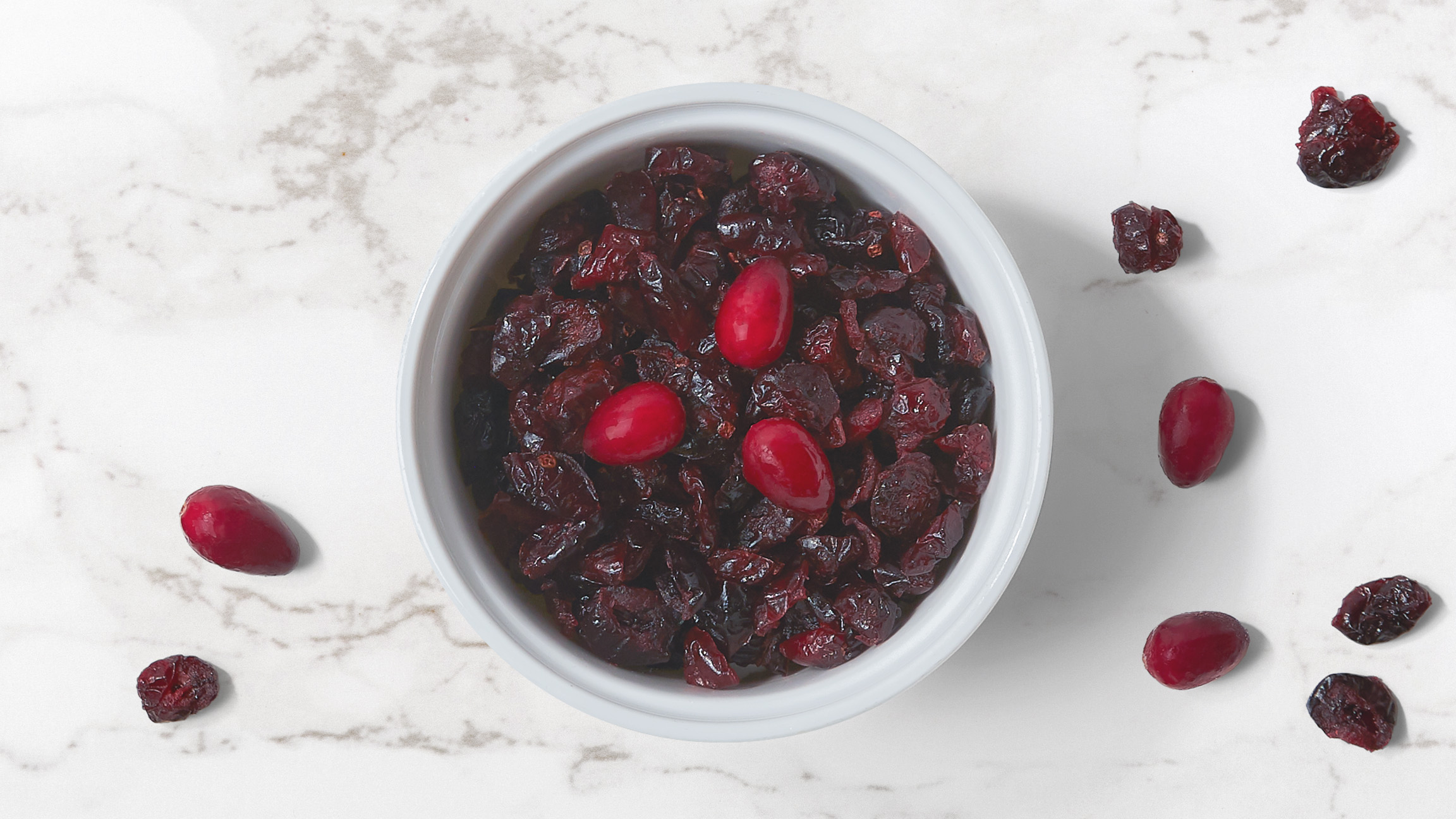 Bowl of dried cranberries in a white bowl with dried and fresh cranberries scattered on a marble counter